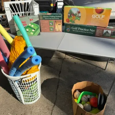 Sale Photo Thumbnail #444: Selection of outdoor recreation equipment, featuring colorful pool noodles in a laundry basket, boxed golf practice nets and putting systems on a folding table, and a box of sports balls.