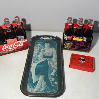 Sale Photo Thumbnail #412: Collection of vintage Coca-Cola memorabilia, including a vintage metal serving tray featuring a 1920s-style woman, two full six-packs of glass Coca-Cola bottles in cardboard cartons, and a small red Coca-Cola box.