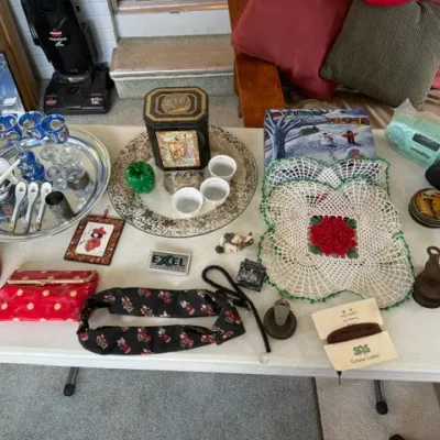 Sale Photo Thumbnail #244: Collection of tabletop estate items including a silver-plated serving tray, ceramic tea cups and spoons, crocheted doilies, vintage tins, a red polka dot wallet, and small decoratives.