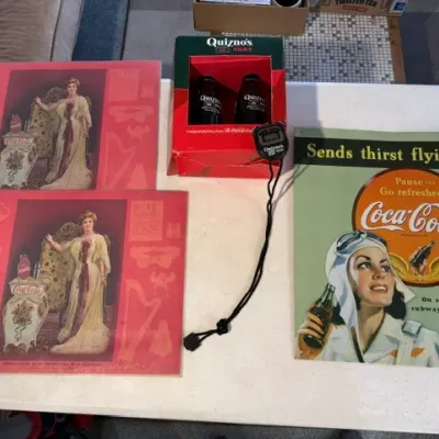 Sale Photo Thumbnail #225: Collection of Coca-Cola vintage-style advertising art prints and tin signs alongside a boxed Quiznos Subs promotional set with a digital stopwatch.