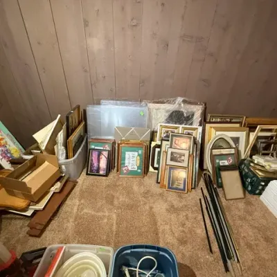 Sale Photo Thumbnail #344: Large assortment of framed artwork, empty picture frames, and wall decor lining a wood-paneled basement room.