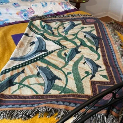 Sale Photo Thumbnail #298: Woven fringe throw blanket featuring a dolphin and seaweed motif, displayed folded on a bed.