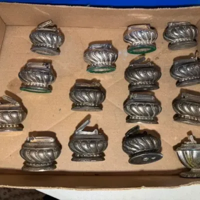 Sale Photo Thumbnail #56: Collection of vintage silver-toned metal table lighters with gadrooned bases, housed together in a cardboard tray.