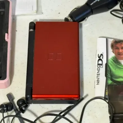 Sale Photo Thumbnail #418: Metallic red Nintendo DS Lite handheld gaming console, shown closed alongside various accessories including styluses, headphones, and a car charger.