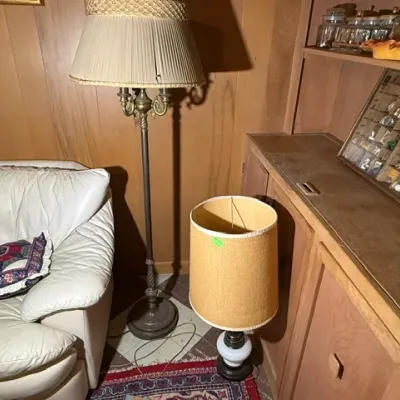 Sale Photo Thumbnail #252: Vintage brass floor lamp with pleated fabric shade and a mid-century table lamp with an oversized textured drum shade, set alongside wood furniture and a white leather sofa.