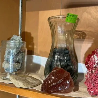 Sale Photo Thumbnail #119: Collection of decorative mineral specimens and coral on a shelf, including a fluted glass vase filled with dark river stones, a jar of grey rocks, a piece of red slag glass, and a large red coral specimen.