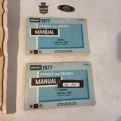 Sale Photo Thumbnail #236: Pair of 1977 GMC owner's and driver's manuals for models 1500 through 3500, shown alongside small metal emblems and a chart.