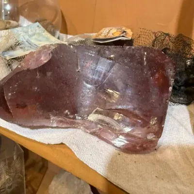 Sale Photo Thumbnail #123: Large chunk of dark red or purple slag glass resting on a wooden shelf alongside decorative stones.