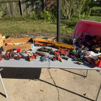 Sale Photo Thumbnail #468: Large collection of vintage toy vehicles, including die-cast cars, plastic fire trucks, semi-trucks, and a pink storage tub full of additional toys displayed on a folding table.