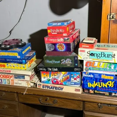 Sale Photo Thumbnail #402: Large collection of vintage board games stacked on a wooden desk, including Monopoly, Clue, Sorry!, Password, and SongBurst.