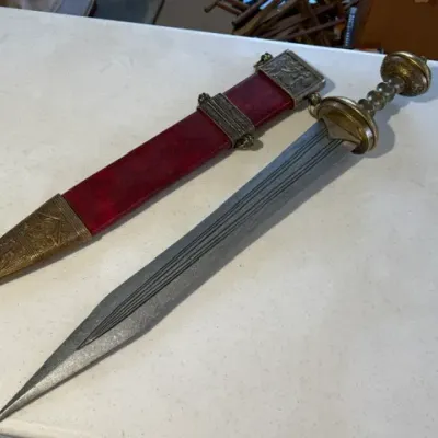 Sale Photo Thumbnail #219: Roman gladius-style short sword replica with double-edged blade, ornate brass hilt, and matching red scabbard with embossed brass fittings.