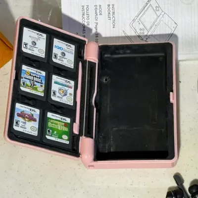 Sale Photo Thumbnail #419: Pink Nintendo DS console carrying case containing six game cartridges, including New Super Mario Bros., alongside a instruction booklet and red Nintendo 3DS system.
