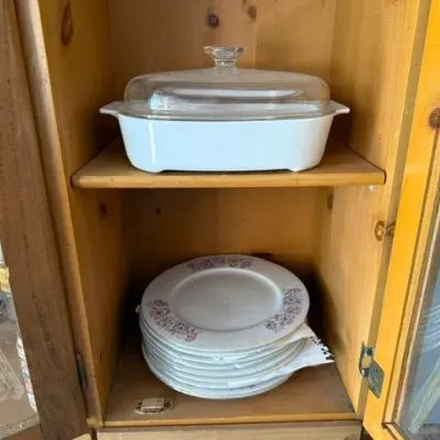 Sale Photo Thumbnail #487: Vintage white Pyrex-style casserole dish with clear glass lid stacked above a set of floral ceramic plates inside a wooden cabinet.