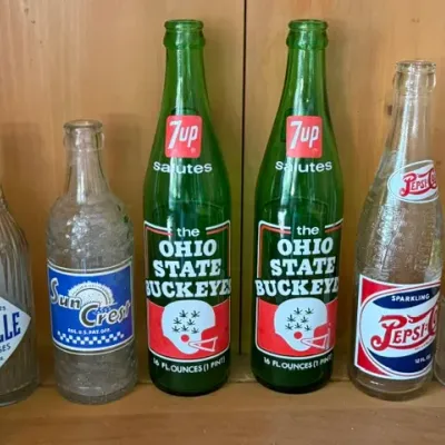 Sale Photo Thumbnail #485: Collection of vintage soda bottles, featuring a pair of green 7Up 16 oz bottles saluting the Ohio State Buckeyes alongside Pepsi-Cola, Sun Crest, and Chille Beverages clear glass bottles.