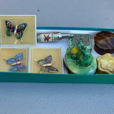 Sale Photo Thumbnail #450: Collection of vintage small collectibles and trinkets in a box, including cloisonné butterfly brooches in display cases, porcelain frog figurines, a miniature Budweiser bottle, and decorative pill boxes.