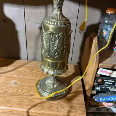 Sale Photo Thumbnail #336: Vintage brass table lamp base with ornate acanthus leaf detailing, displayed next to a wooden shelf holding VHS tapes.