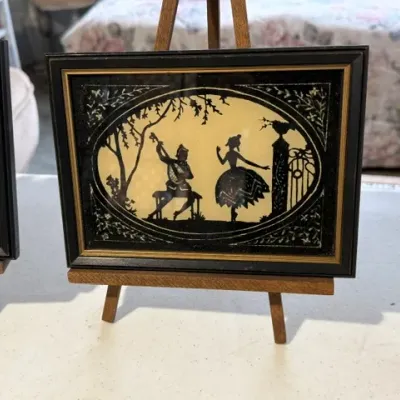 Sale Photo Thumbnail #194: Vintage reverse glass silhouette artwork depicting a seated musician and a dancing figure, displayed in a black frame with gold inner trim on a small wooden easel.