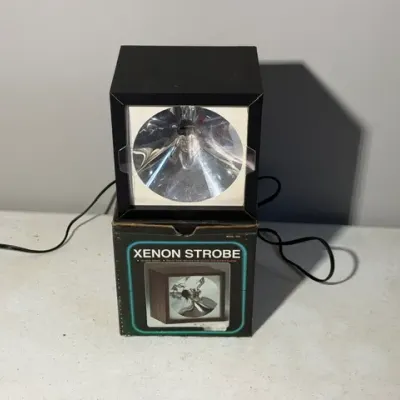 Sale Photo Thumbnail #415: Vintage Xenon Strobe light in a black cube housing, resting on its original retail box.