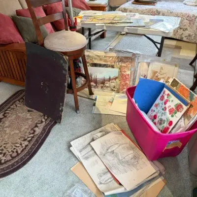 Sale Photo Thumbnail #195: Collection of framed artwork, prints, and sketches spread across a room, alongside a wooden bar stool, Mission-style sofa, and storage totes filled with art supplies.