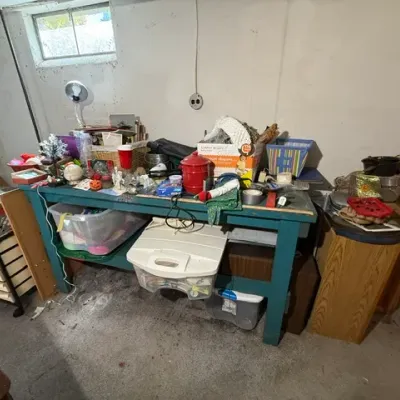 Sale Photo Thumbnail #370: Teal painted wooden workbench filled with various household knick-knacks, plastic storage bins, crafts, and tools in a basement setting.