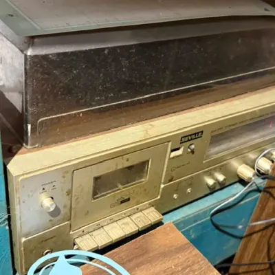 Sale Photo Thumbnail #372: Vintage Seville AM/FM stereo receiver with a built-in cassette tape deck and top-mounted turntable cover, showing visible dust and surface wear.