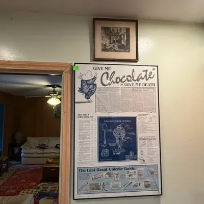 Sale Photo Thumbnail #309: Framed novelty 'Give Me Chocolate or Give Me Death' poster alongside a small framed architectural print mounted on the wall.