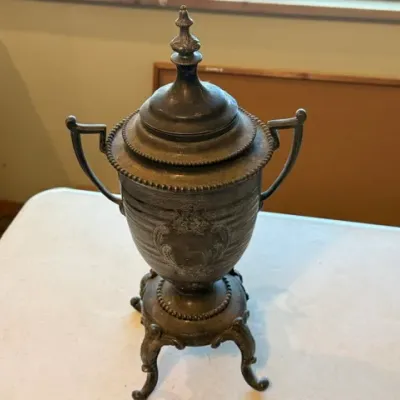 Sale Photo Thumbnail #39: Tarnished silver-plated or pewter urn with a lid, featuring ornate engraving, dual handles, and a four-footed pedestal base.