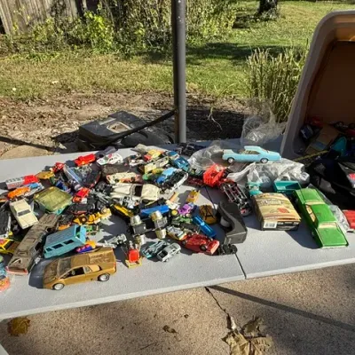 Sale Photo Thumbnail #477: Large collection of vintage die-cast toy cars, plastic models, and vehicles arranged on a folding outdoor table.