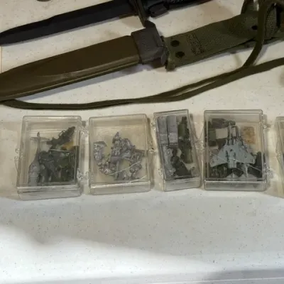 Sale Photo Thumbnail #426: Collection of miniature metal military figures and aircraft in clear plastic display cases, displayed alongside a military-style bayonet knife with sheath.