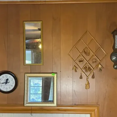 Sale Photo Thumbnail #257: Collection of vintage wall decor mounted on wood paneling, including framed mirrors, an analog wall clock, a decorative straw mobile, and a wooden barometer.