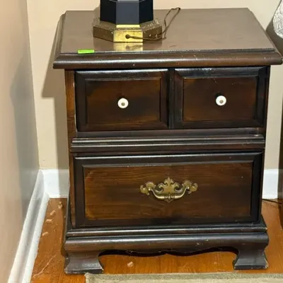 Sale Photo Thumbnail #146: Traditional dark wood nightstand with bracket feet, featuring two upper faux drawers with white knobs and a single deep lower drawer with an ornate brass pull.