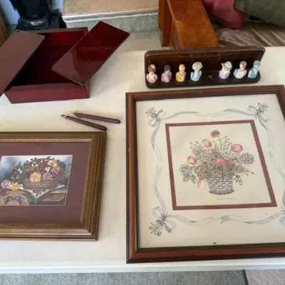 Sale Photo Thumbnail #247: Collection of home decor items featuring two framed floral prints, a red lacquered keepsake box, pair of taper candles, and a vintage set of small porcelain figure thimbles on a wooden rack.