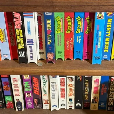 Sale Photo Thumbnail #337: Collection of vintage 1990s VHS tapes lined up on a wooden shelf, including titles like Home Alone, Teenage Mutant Ninja Turtles, Forrest Gump, and Jurassic Park.