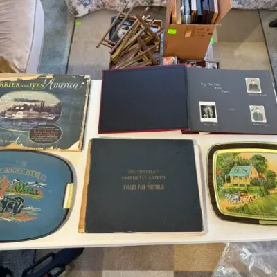 Sale Photo Thumbnail #222: Collection of vintage books and souvenir metal serving trays displayed on a white folding table. Items include a 'Currier and Ives America' book, an antique photo album, and a Great Smoky Mountains tray.