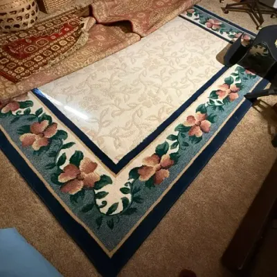 Sale Photo Thumbnail #353: Cream and blue floral area rug with a dark border, displayed alongside additional folded oriental-style rugs.