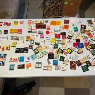 Sale Photo Thumbnail #214: Large vintage matchbook collection featuring various advertising, hotel, and restaurant designs laid out on a white folding table.