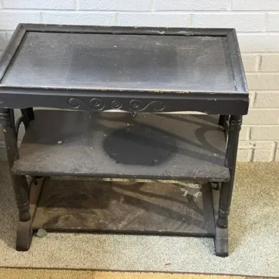 Sale Photo Thumbnail #175: Vintage dark-painted wooden tea cart or utility table featuring carved scroll details and two lower shelves, showing finish wear and dust.