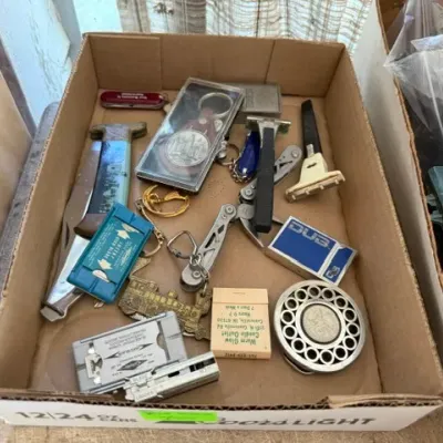 Sale Photo Thumbnail #460: Cardboard box containing a variety of small vintage collectibles and tools, including pocket knives, multi-tools, razor blade dispensers, and keychains.