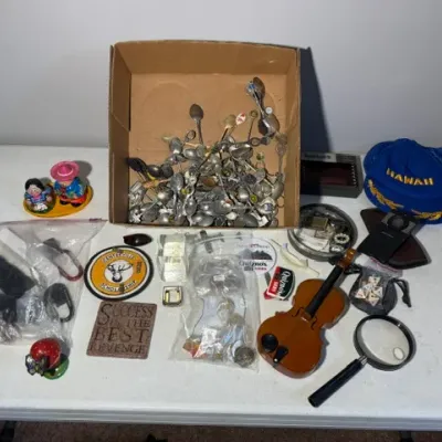 Sale Photo Thumbnail #413: Collection of eclectic estate items including a box of souvenir spoons, miniature wooden violin, blue Hawaii hat, magnifying glass, and small collectibles.
