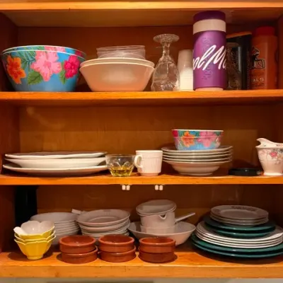 Sale Photo Thumbnail #317: Assorted vintage and modern kitchenware, including ceramic dishware, floral mixing bowls, glass stemware, and small baking ramekins, arranged on wooden cabinet shelves.