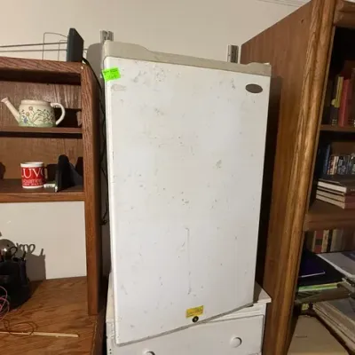 Sale Photo Thumbnail #360: Compact white mini refrigerator positioned between wooden bookshelves, showing light exterior scuffs and wear.