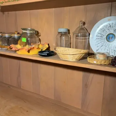 Sale Photo Thumbnail #267: Collection of vintage glass storage jars, small woven baskets, and a commemorative presidential seal signatures plate arranged on wood shelving.