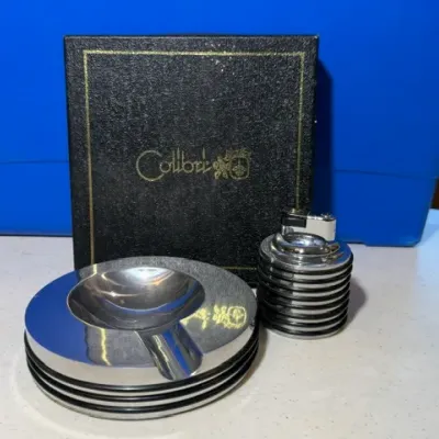 Sale Photo Thumbnail #58: Vintage Colibri chrome table lighter and matching ashtray set with original textured black box, featuring a ribbed mid-century modern design.