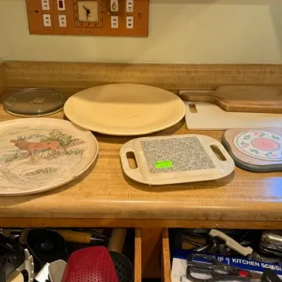 Sale Photo Thumbnail #316: Collection of kitchen items including a yellow Fiesta platter, wooden cutting boards, trivets, and a decorated serving tray.