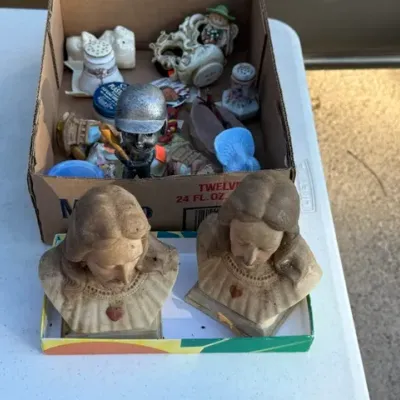 Sale Photo Thumbnail #454: Pair of vintage plaster busts of Jesus and Mary alongside a cardboard box filled with assorted knick-knacks and figurines.
