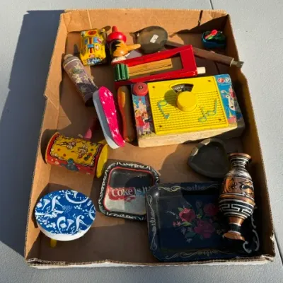 Sale Photo Thumbnail #452: Cardboard flat filled with vintage toys and small collectibles, including a Fisher-Price music box, tin noise makers, small trays, and decorative trinkets.