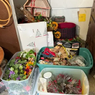Sale Photo Thumbnail #375: Collection of storage tubs containing artificial flowers, holiday wreaths, and crafting supplies, alongside wooden furniture and embroidery hoops.