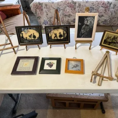 Sale Photo Thumbnail #193: Collection of small framed artwork, silhouette art, cat portraits, and tabletop display easels arranged on a folding table.
