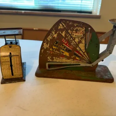 Sale Photo Thumbnail #34: Two vintage metal scales, including a small Pelouze postal scale and a larger egg scale, both showing heavy rust and age-related patina.
