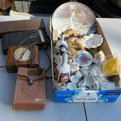Sale Photo Thumbnail #458: Assortment of vintage collectibles, including a Zenith transistor radio in leather case, folding Kodak camera, small wooden desk clock, ceramic bells, mini tea set pieces, and a moon landing commemorative plate.